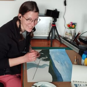 Annika sitting at her studio desk drawing and listening to music and smiling at the camera.