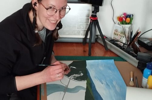 Annika sitting at her studio desk drawing and listening to music and smiling at the camera.