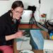 Annika sitting at her studio desk drawing and listening to music and smiling at the camera.