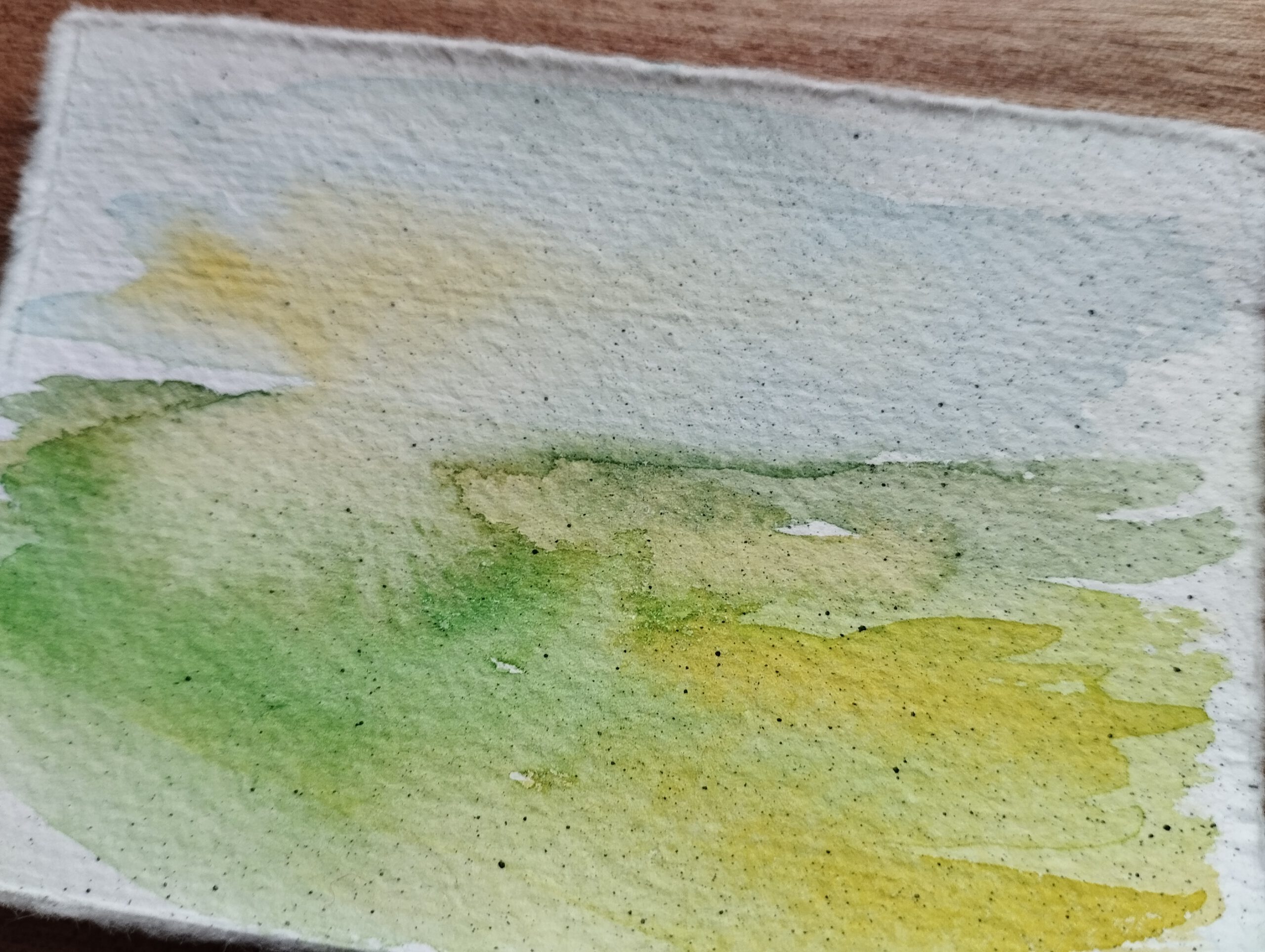 Landschaft in Aquarell auf Büttenpapier. Auf der unteren linken Hälfte ist eine grüne Wiese zu sehen, die nach rechts in Gelbtöne übergeht. Im Himmel scheint links durch die Wolken diffuses Sonnenlicht.