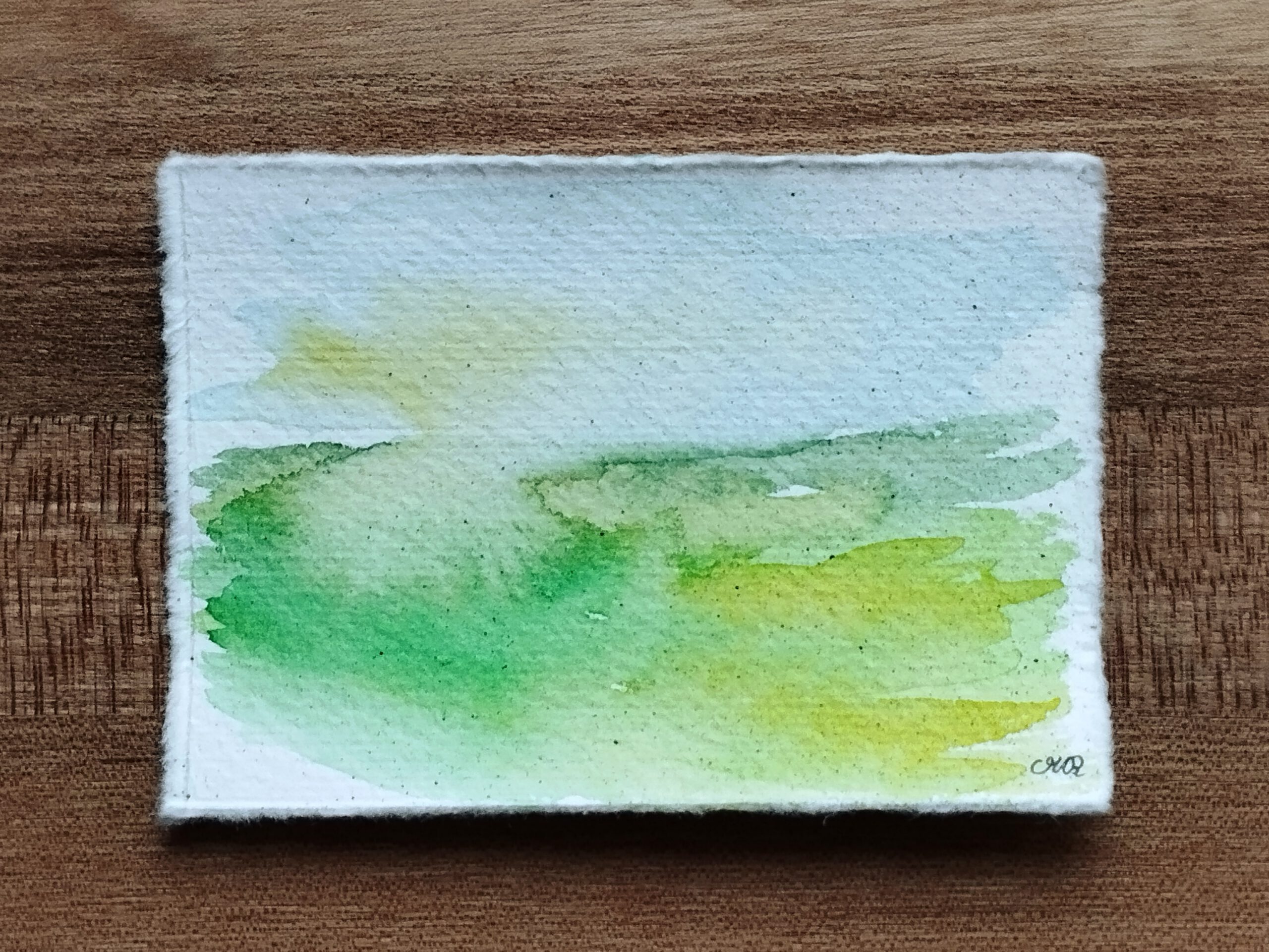 Landschaft in Aquarell auf Büttenpapier. Auf der unteren linken Hälfte ist eine grüne Wiese zu sehen, die nach rechts in Gelbtöne übergeht. Im Himmel scheint links durch die Wolken diffuses Sonnenlicht.