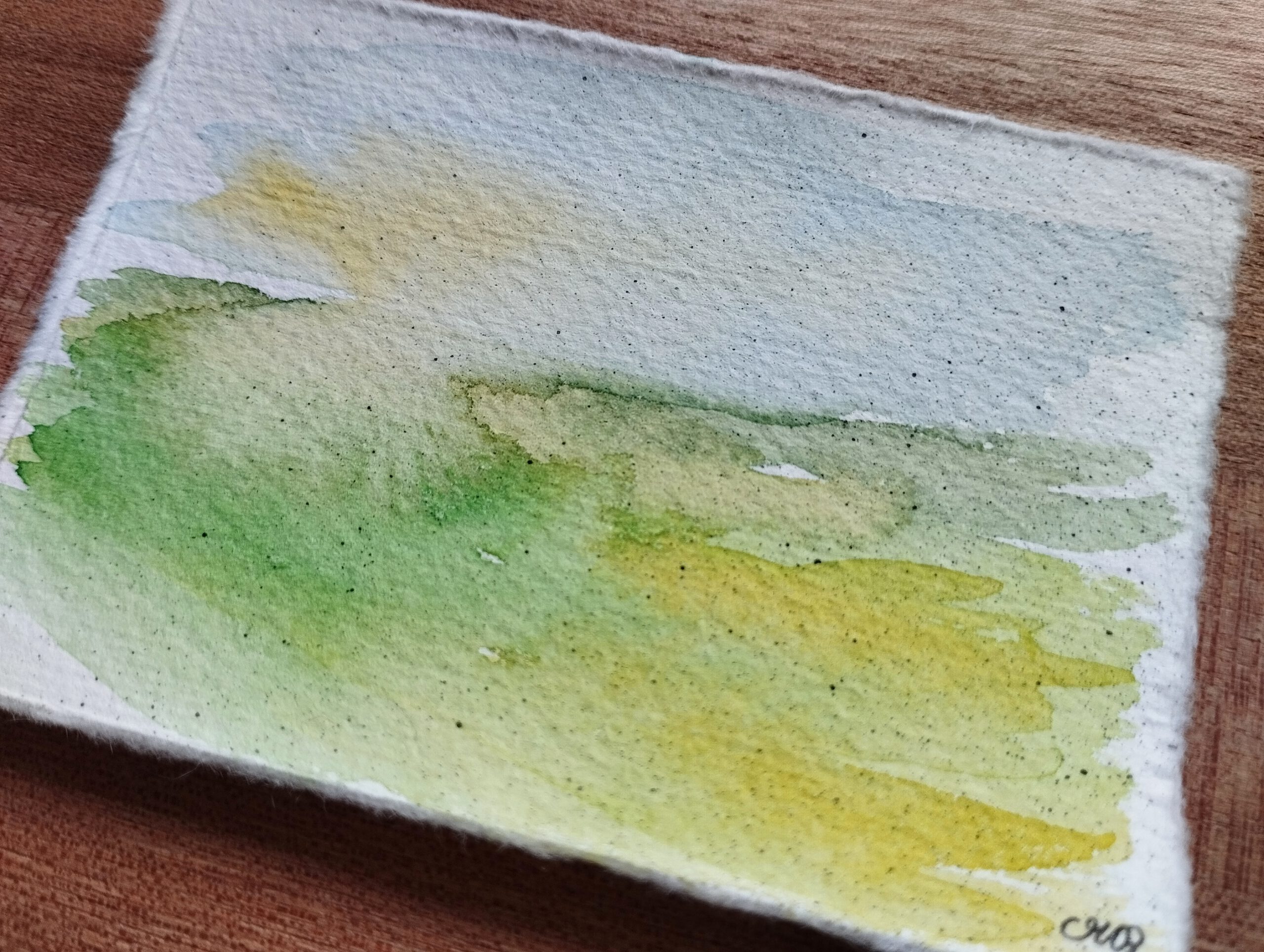Landschaft in Aquarell auf Büttenpapier. Auf der unteren linken Hälfte ist eine grüne Wiese zu sehen, die nach rechts in Gelbtöne übergeht. Im Himmel scheint links durch die Wolken diffuses Sonnenlicht.