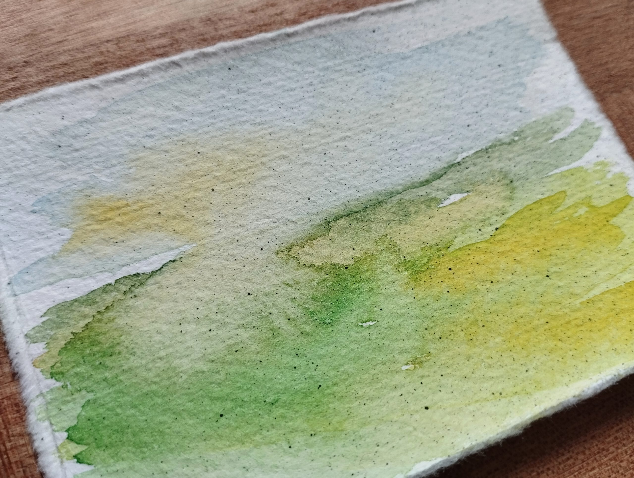 Landschaft in Aquarell auf Büttenpapier. Auf der unteren linken Hälfte ist eine grüne Wiese zu sehen, die nach rechts in Gelbtöne übergeht. Im Himmel scheint links durch die Wolken diffuses Sonnenlicht.