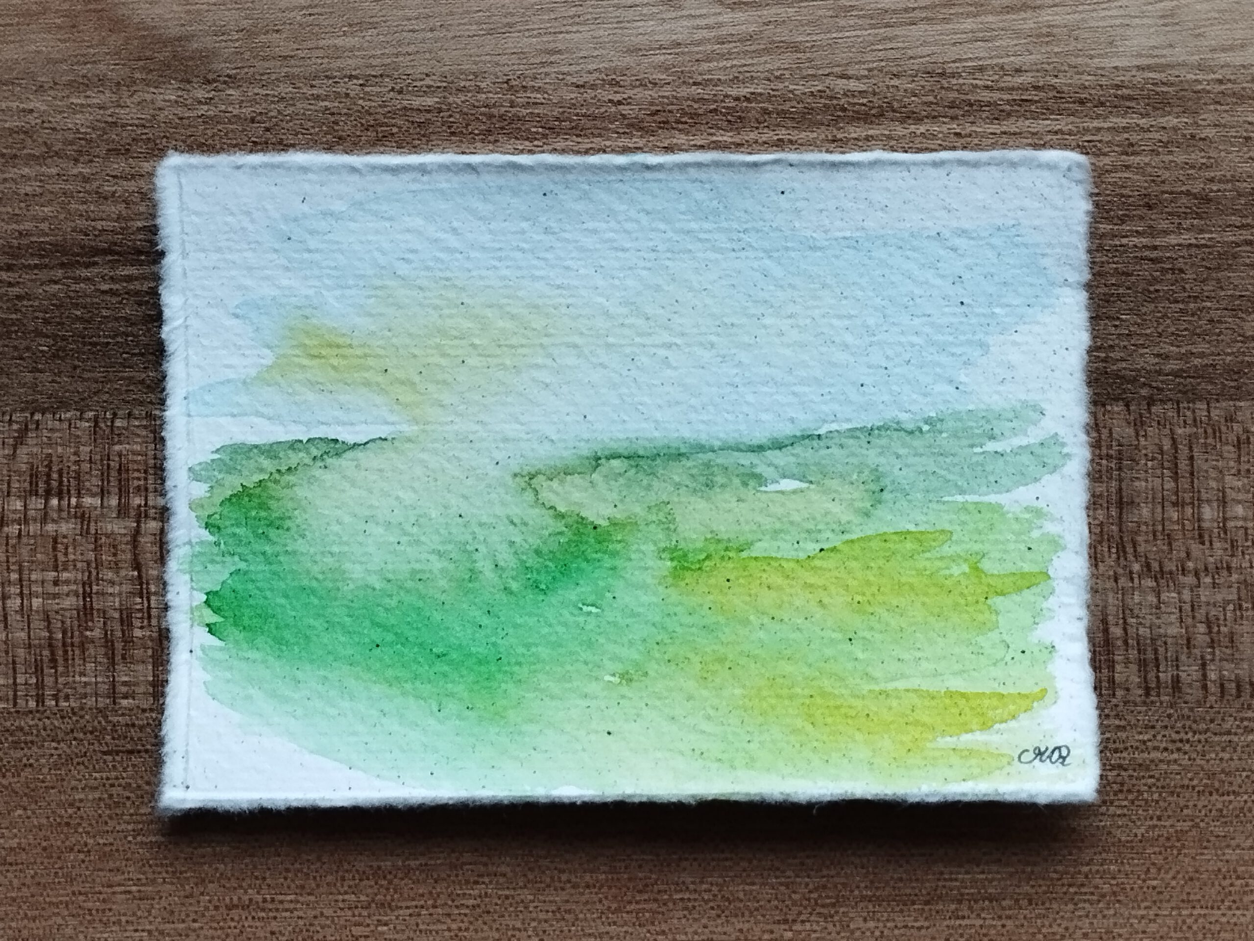 Landschaft in Aquarell auf Büttenpapier. Auf der unteren linken Hälfte ist eine grüne Wiese zu sehen, die nach rechts in Gelbtöne übergeht. Im Himmel scheint links durch die Wolken diffuses Sonnenlicht.