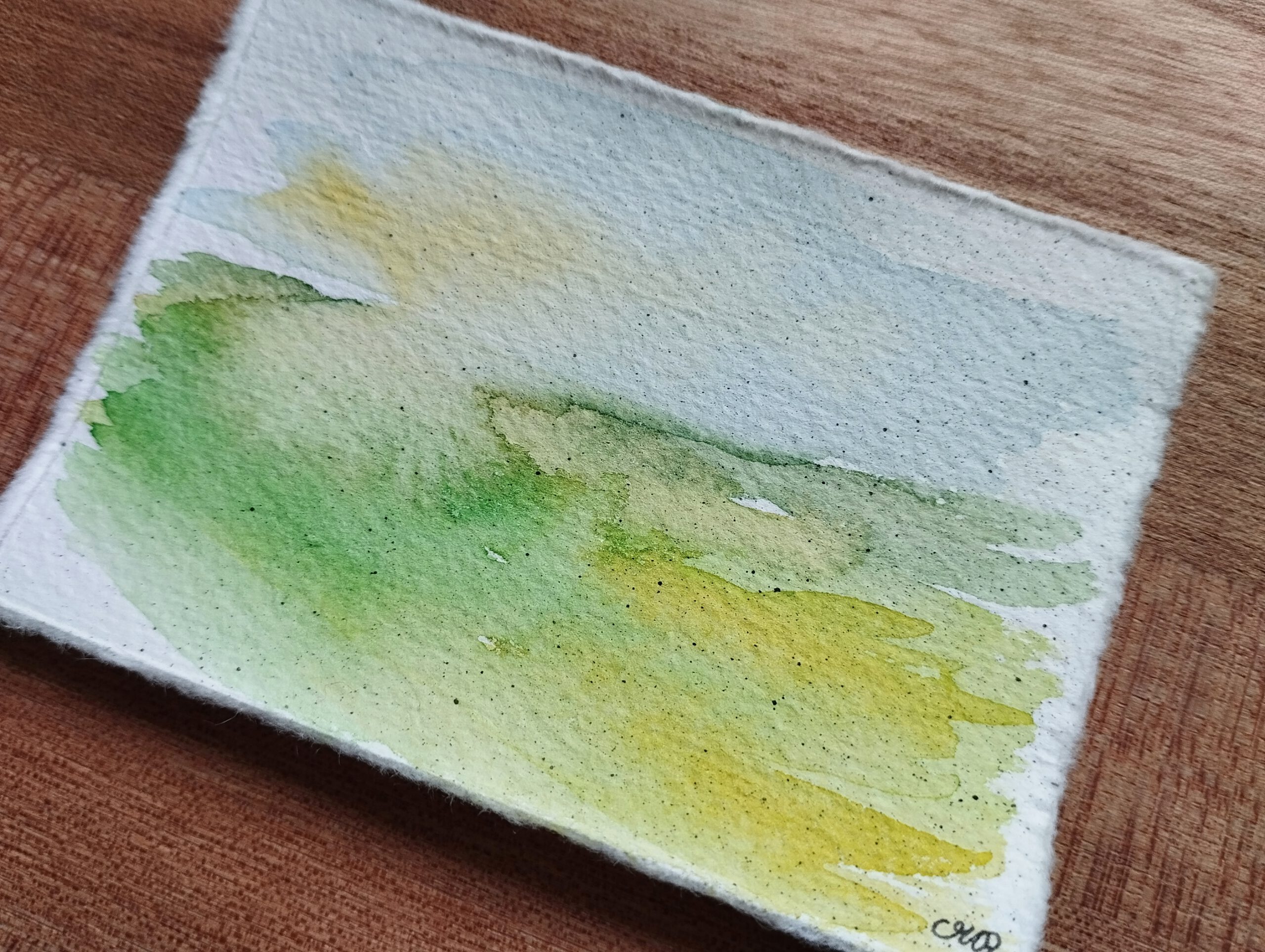 Landschaft in Aquarell auf Büttenpapier. Auf der unteren linken Hälfte ist eine grüne Wiese zu sehen, die nach rechts in Gelbtöne übergeht. Im Himmel scheint links durch die Wolken diffuses Sonnenlicht.