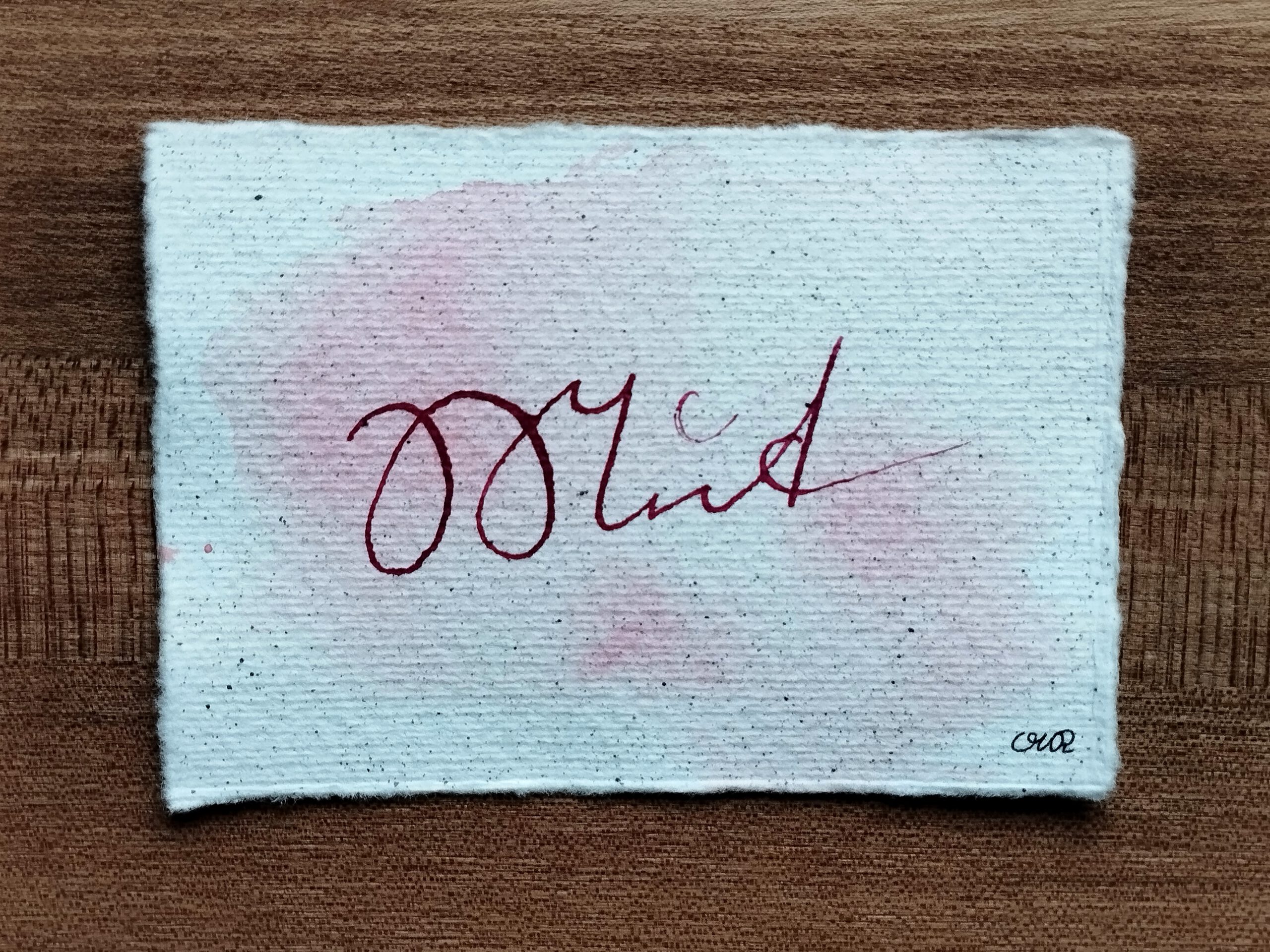 Schriftzug "Mut" in Kurrentschrift in magentafarbener Tusche auf Büttenpapier mit Tintensprenkeln. Im Hintergrund rot-pinke Auqarellfarbe.