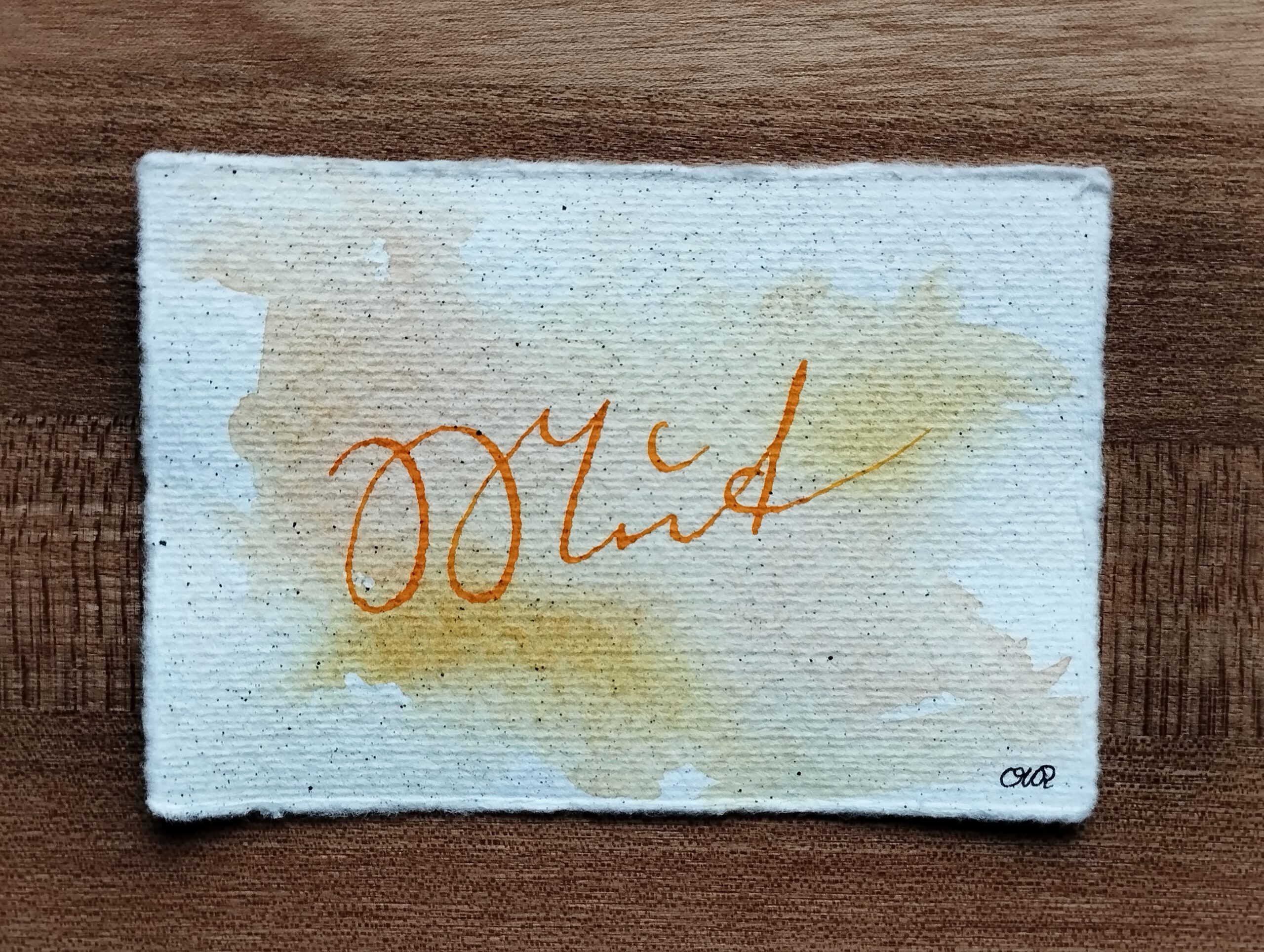 Schriftzug "Mut" in Kurrentschrift in goldgelber Tusche auf Büttenpapier mit Tintensprenkeln. Im Hintergrund gelb-rote Auqarellfarbe.