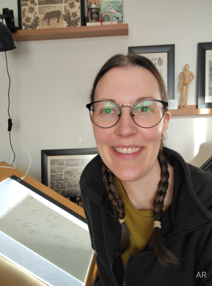 Selfie von Annika Ruhwedel in ihrem Studio. Sie sitzt auf ihrem Stuhl vor dem Zeichenbrett und dreht sich mit Gesicht und Oberkörper zur Kamera. Auf dem beleuchteten Zeichenbrett liegt ein Papier, auf dem verschiedene Schriftzüge erkennbar sind.