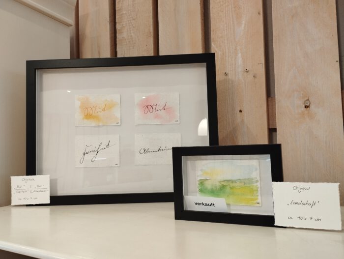 Vier A7-Originale mit Schriftzügen in einem größeren Rahmen. Daneben ein einzelnes gerahmtes A7-Original mit einem Landschafts-Aquarell, an dem ein "verkauft"-Schildchen hängt.