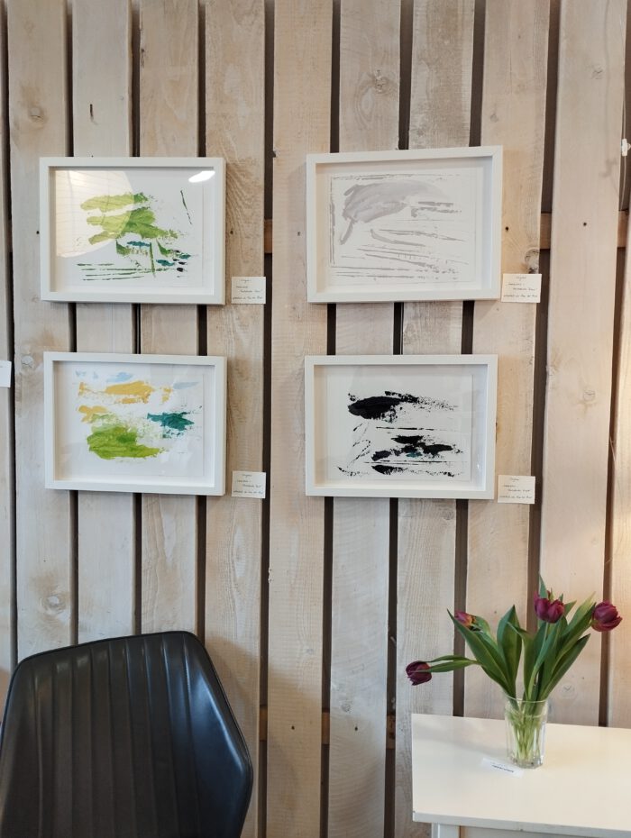 Vier "Werrablick"-Farbstudien hängen als Gruppe an der Wand der Brot-Galerie. Rechts darunter steht ein weißes Tischchen mit einem Strauß lilafarbener Tulpen. Links daneben ein schwarzer Stuhl.
