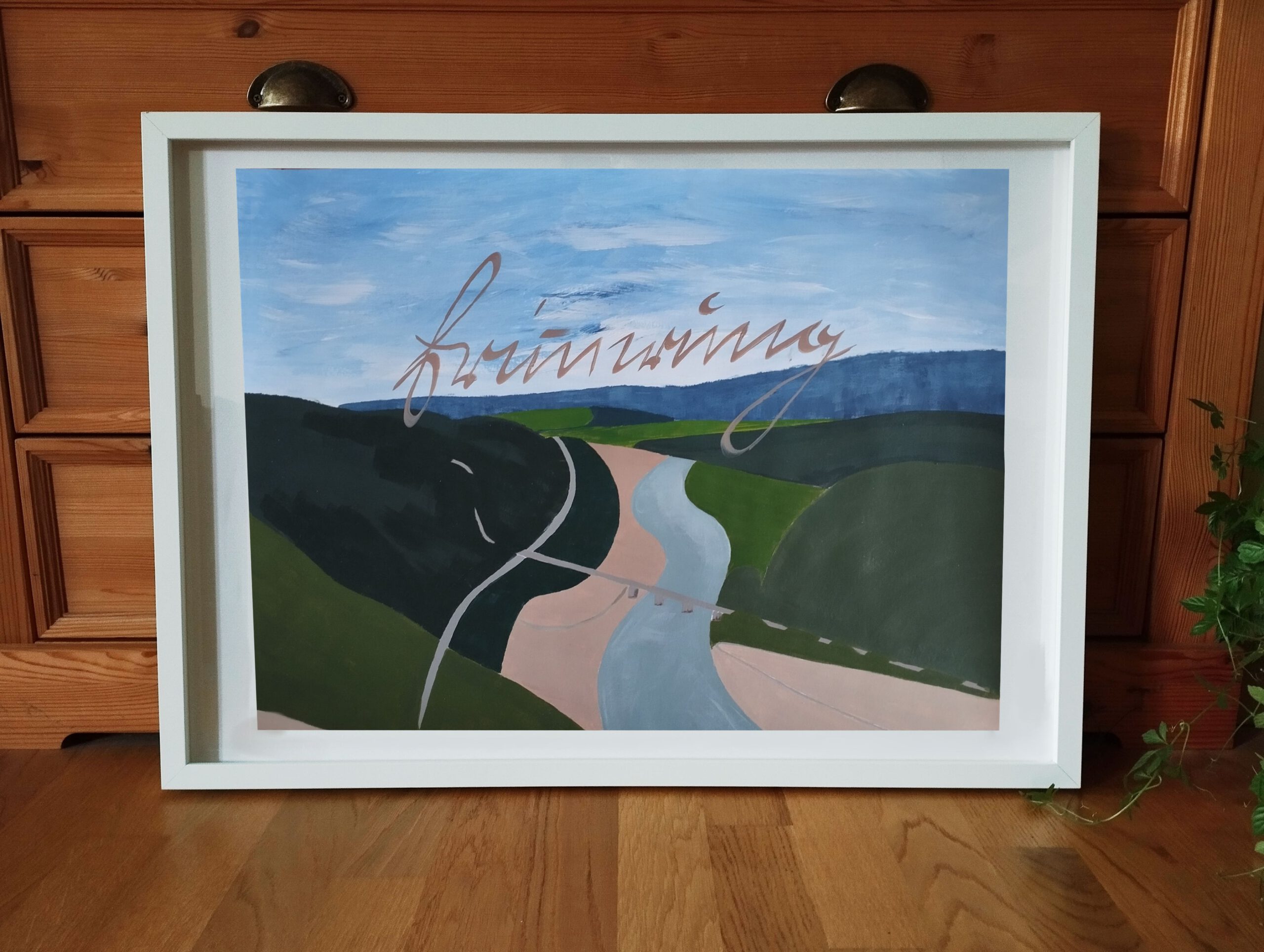 Zu sehen ist ein in einem weißen Rahmen gerahmtes Bild: Eine hügelige Landschaft aus der Vogelperspektive in Grün-, Blau- und Beigetönen. In der Mitte schlängelt sich ein Fluss durchs Bild, den eine Brücke überspannt. Im oberen Teil des Bildes steht leicht schräg von links unten nach rechts oben "Erinnerung" in beiger Kurrentschrift. Durch die Vogelperspektive entsteht ein Gefühl von Weite. Die Arbeit steht auf einem Parkettboden, angelehnt an eine Vitrine.